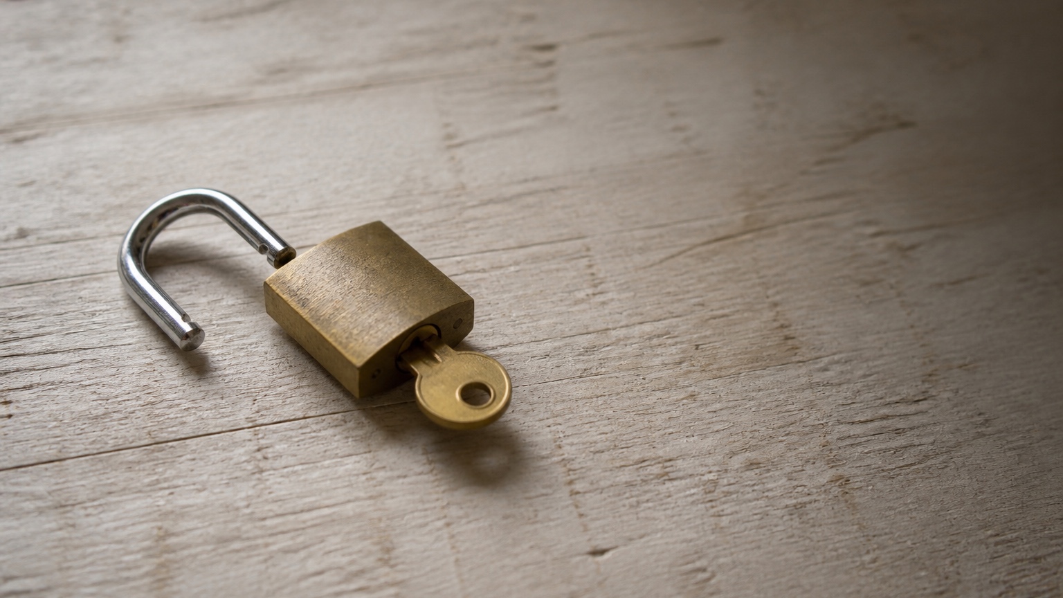 A brass padlock lying open on pale weathered wood, its steel shackle swung wide to the left and a brass key still sitting in the keyhole, with bare wood filling the right half of the frame